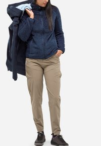 Femme portant une veste en polaire bleue marine et un pantalon beige, tenant un imperméable bleu marine sur son épaule, se tenant avec une main dans la poche.