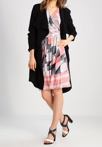 Czarny długi płaszcz na wzorzystej sukience z różowymi, białymi i czarnymi geometrycznymi wzorami. Modelka ma na sobie czarno-białe sandały na obcasie.