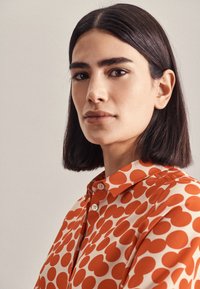 Blusa a maniche corte color crema con grandi pois arancioni, dotata di colletto abbottonato e tessuto liscio e leggero.