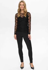 Čierny vrstvený outfit pozostávajúci z priesvitnej blúzky s dlhým rukávom a textúrovaným dizajnom, pod ktorou je čierny tielko, doplnený o slim-fit čierne nohavice a podpätky.