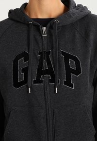 Sweat à capuche zippé gris foncé avec un grand logo noir "GAP" sur la poitrine, doté de cordons de serrage, de poches avant et d'une texture douce.