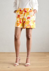 Pantaloni corti floreali in tonalità giallo, arancione e rosso su uno sfondo crema, dotati di tasche laterali e una texture di tessuto liscia.