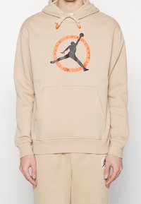 Felpa beige con tasca a marsupio anteriore, caratterizzata da un logo Jumpman nero e un design circolare arancione. Mischia di cotone, vestibilità regolare.