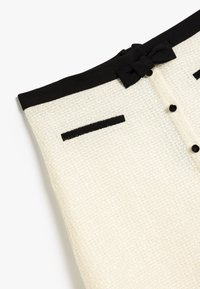 Weiße Tweed-Rock mit schwarzem Bund, kleinen Schleifendetails und schwarzen Akzenten, zwei falschen Taschen und Knöpfen. Strukturierter Stoff.