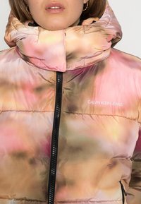 Giacca imbottita con un motivo a gradiente rosa e beige. Presenta un colletto alto, chiusura con zip e logo ricamato "CALVIN KLEIN JEANS".