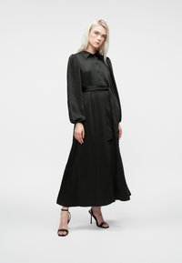 Naine, kellel on pikad blondid juuksed, kannab pikka musta nööbiga kleiti, millel on puhvitud varrukad, ja musti kõrge kontsaga sandaale, seistes Valgel taustal.