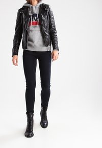 Svart lädermotorcykeljacka över en grå huvtröja med röd och blå logotyp, i kombination med svarta skinny jeans och snörda combat boots.