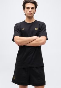 Νεαρός άνδρας με σταυρωμένα τα χέρια, φοράει μαύρο t-shirt Adidas και μαύρα σορτς δίχτυ με χρυσά λογότυπα FC Bayern Munich.