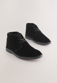 Paar schwarze Wildleder-Stiefeletten mit Schnürsenkeln und niedrigen Gummisohlen auf hellem Hintergrund.