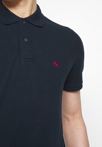 Muž ve černém polokošili s krátkým rukávem, s dvěma knoflíky a malým červeným logem losa na levé části hrudi.