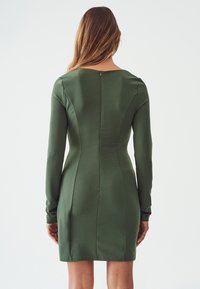 Robe ajustée verte olive à manches longues, avec des coutures discrètes et une fermeture éclair au dos. Texture lisse et coupe midi.