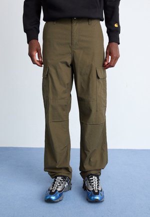 Personne portant un pantalon cargo vert olive et des baskets noires avec des détails bleus et argentés, debout sur un sol bleu clair.