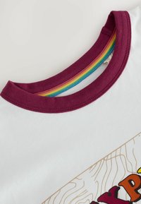 Bílý tričko s vínovým límečkem a mnohobarevným pruhovaným vnitřním výstřihem; detaily zahrnují grafický design v hnědých tónech.