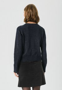 Pulover texturat de culoare bleumarin, cu decolteu rotund și mâneci lungi, asortat cu o fustă neagră. Materialul are un efect delicat de strălucire.