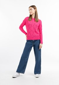 Maglione lavorato a maglia di un vivace colore rosa con motivi a diamante abbinato a jeans a gamba larga blu e sneakers in tela bianche. Modella posa su uno sfondo bianco.