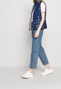 Marinefarbener Steppwesten mit hohem Kragen, kombiniert mit einem hellrosa Hemd und hellblauen Jeans. Weiße Sneakers runden das Outfit ab.