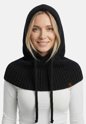 Capa con capucha negra de punto con textura acanalada, con cordones y un detalle de cuero. Llevada sobre una camiseta blanca de manga larga.