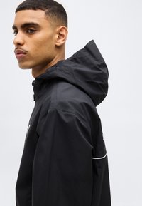 Jeune homme aux cheveux courts portant une veste à capuche noire, faisant face à gauche sur un fond clair neutre.