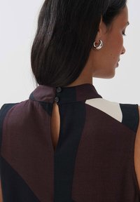 Ärmellose Bluse aus dunkelburgunderfarbenem und schwarzem Stoff mit einem Schlüsselloch-Ausschnitt hinten, ausgestattet mit zwei schwarzen Knöpfen am Ausschnitt und geometrischen Designelementen.