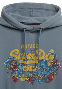 Sudadera con capucha gris que presenta un gráfico bordado en azul y rojo con una cara de demonio, acentos florales y un logotipo vintage en amarillo.
