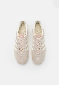 Beige suede sneakers with white stripes, featuring a lace-up design, light pink accents on the tongue, and a patterned insole.