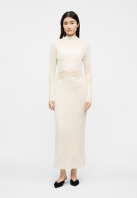 Femme portant une robe midi crème à manches longues côtelées avec une ceinture à la taille, debout devant un fond blanc uni en chaussures plates noires pointues.