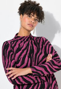 Svart klänning med rosa zebraränder, långa ärmar, hög halsringning, mjukt tyg som har ett texturerat mönster. Modellen har hängande örhängen.