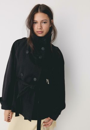 Jeune femme aux longs cheveux foncés portant un manteau noir boutonné avec un col montant et une ceinture, se tenant devant un fond blanc uni.