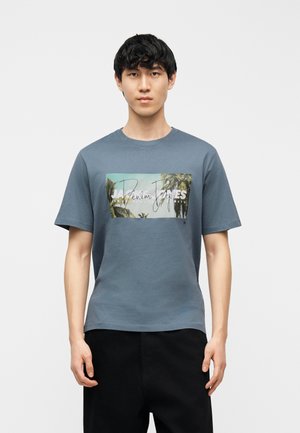 Jeune homme debout, portant un t-shirt bleu avec un graphique rectangulaire de palmiers et du texte, associé à un pantalon noir sur un fond uni.