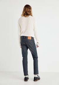 Lys beige, ribbet strikkegenser, kombinert med mørke, tettsittende jeans med en rød og gul etikett på baklivet og oppbrettede ermer.