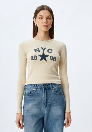 Beige langekleurige t-shirt met lange mouwen en ribbeltextuur, met "NYC" en een ster geborduurd in marineblauw, gestyled met high-waisted blauwe denim jeans.