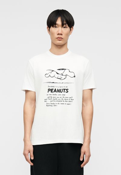 Νέος άνδρας που φοράει λευκό T-shirt με ασπρόμαυρο γραφικό του Snoopy που τρέχει και κείμενο από το κόμικ Peanuts στο εμπρόσθιο μέρος.