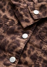 Tissu en velours côtelé brun à taches foncées avec des boutons-pression argentés et des coutures visibles sur la manche et la poche d'une veste.