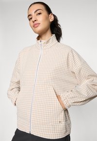 Lightweight beige and white checkered jacket with a high collar, zip front, and two front pockets. Elastic cuffs and loose fit design.