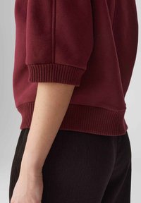 Felpa borgogna con polsini e orlo a coste. Tessuto morbido con vestibilità comoda. Indossata con pantaloni neri testurizzati.