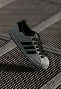Black and gray sneaker with a leather upper, light gray sole, black laces, and three black stripes. Textured toe cap with logo detail.