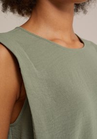 Ramiona i dekolt osoby ubranej w bezrękawnik z lekko zielonego, teksturowanego materiału na neutralnym tle.