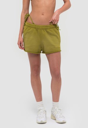 Person iført olivengrønne shorts med elastisk talje, hvide sokker og hvide sneakers, stående mod en ensfarvet lys baggrund.