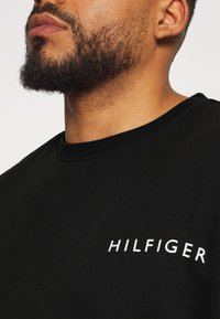Homme avec une barbe taillée portant un t-shirt noir à col rond avec le texte blanc « HILFIGER » sur la poitrine sur un fond uni.