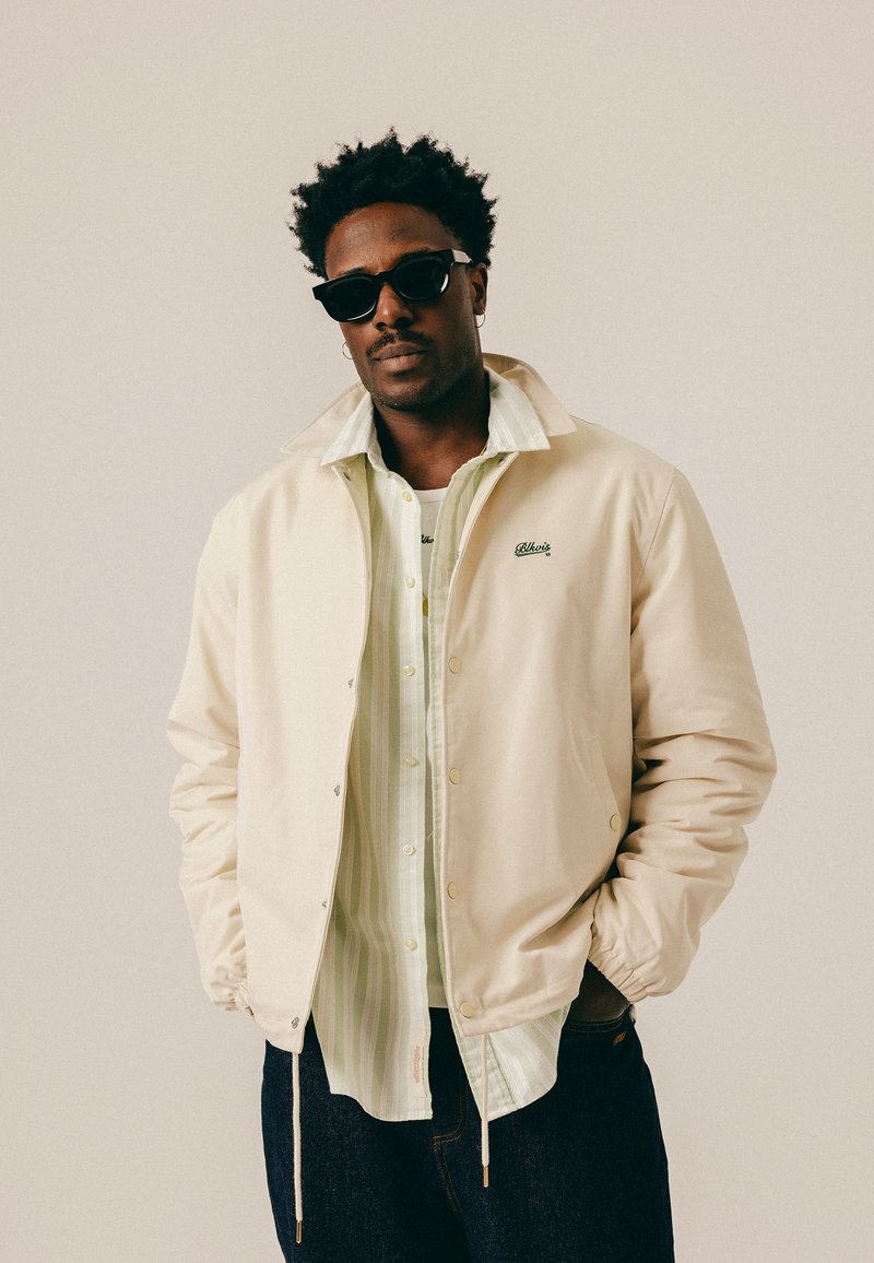 Homme portant des lunettes de soleil noires, une veste crème, une chemise à rayures claires et un jean foncé, debout les mains dans les poches devant un fond uni.