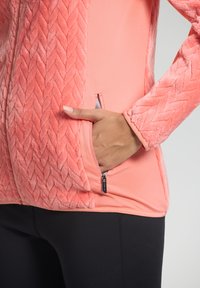 Hand in de met rits sluitende zak van een koraalroze, gestructureerde fleece jas, gedragen met zwarte leggings op een neutrale achtergrond.
