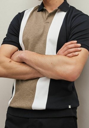 Man wearing short-sleeve black polo with wide beige and white vertical stripes, arms crossed over chest against plain background.