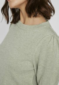 Pull léger vert en tissu tricoté doux. Présente un col rond et des détails de manches bouffantes. Texture lisse.