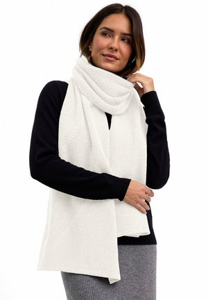 Frau trägt schwarzen Pullover und grauen Rock, richtet einen großen weißen gestrickten Schal, der um ihren Hals und ihre Schultern gewickelt ist, zurecht.