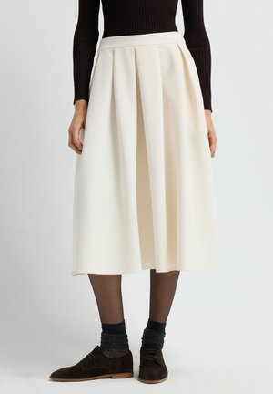 Donna che indossa una gonna midi plissettata color crema con collant neri trasparenti, calzini luccicanti scuri, scarpe stringate marroni e un top a maniche lunghe a coste nere.