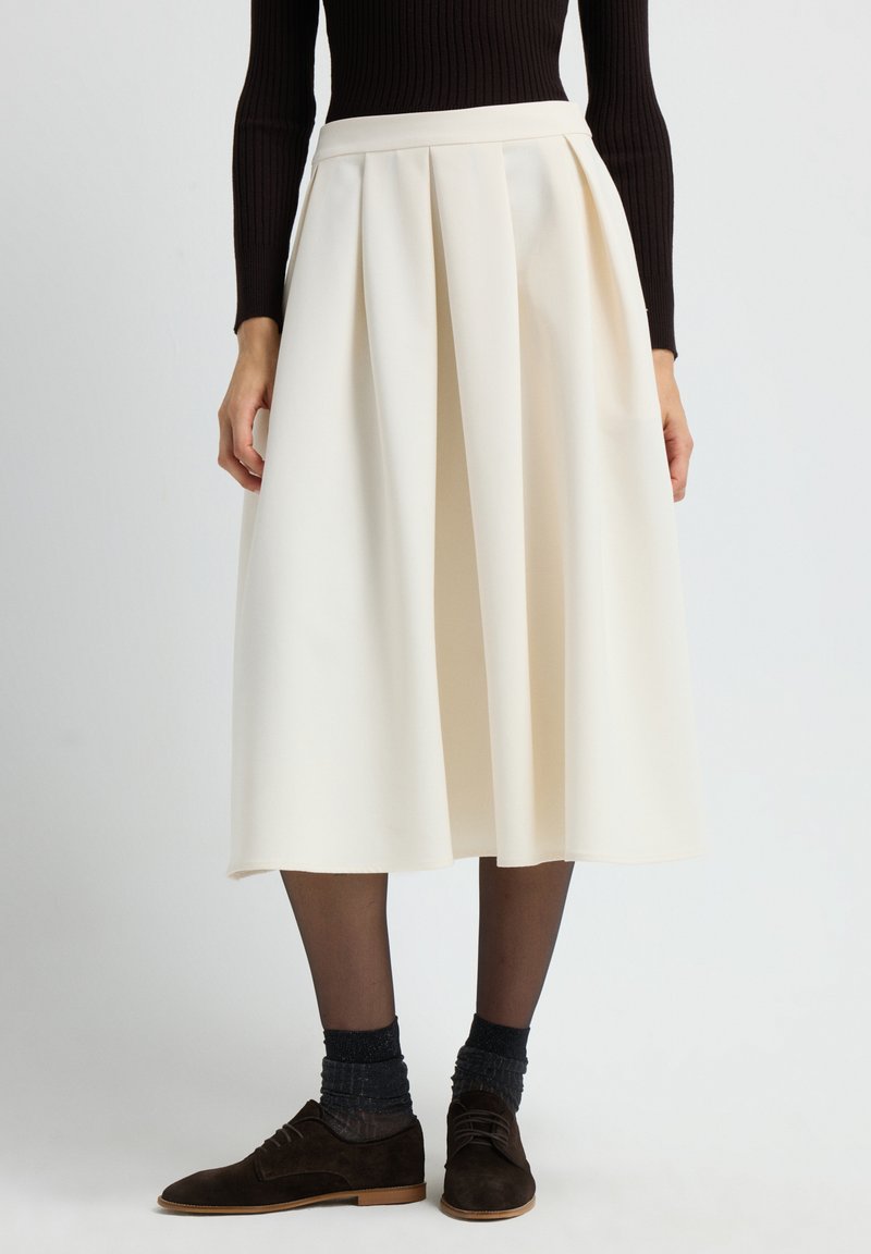 Femme portant une jupe midi plissée crème avec des collants transparents noirs, des chaussettes scintillantes sombres, des chaussures à lacets brunes et un haut noir à manches longues côtelé.