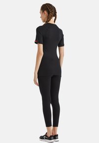 Svart atletisk topp med korta ärmar och åtsittande design, i kombination med svarta leggings och sneakers, som visar en slät yta. Bakifrån visad.