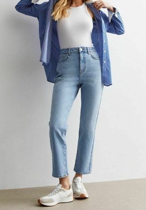 Kobieta ubrana w jasnoniebieskie jeansy z wysokim stanem, biały top na ramiączkach, niebieską koszulę w paski noszoną na rozpięto oraz białe trampki, stojąca na tle gładkiej ściany.