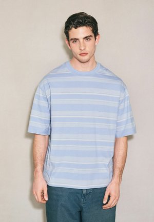 Giovane uomo con capelli corti e scuri indossa una camicia a righe azzurro chiaro a maniche corte e pantaloni scuri, in piedi davanti a uno sfondo chiaro e semplice.