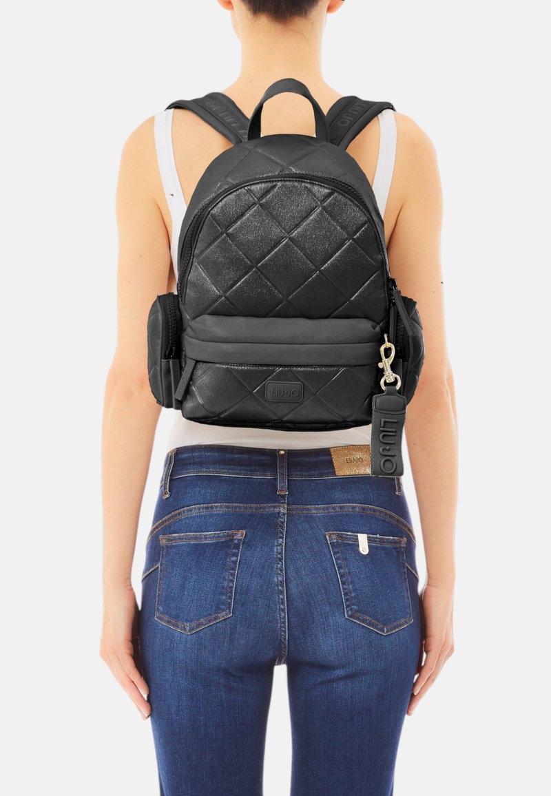 Zaino nero trapuntato con finitura texturizzata, doppie spalline, tasca frontale con zip e etichetta con logo. Indossato sopra una maglietta bianca e jeans.